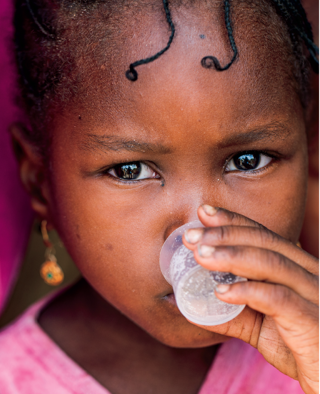 A young girl drinking from a small cup, wearing earrings and a pink garment.