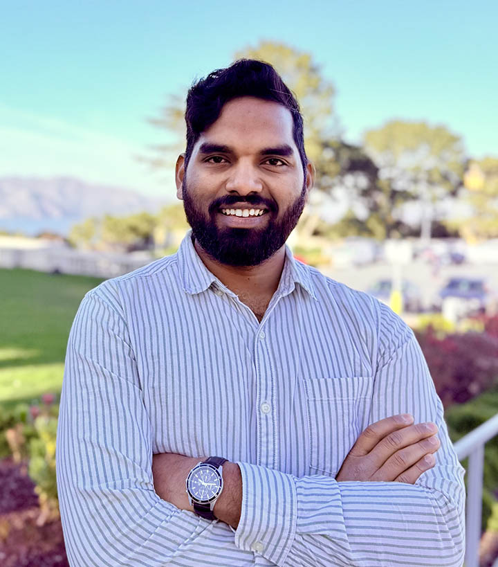 Smiling man with arms crossed outdoors near green grass.