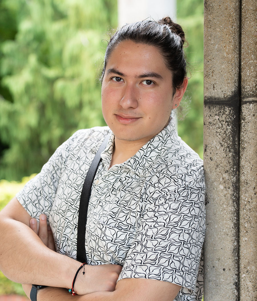 A young man with long hair posing confidently near a stone structure, surrounded by greenery. Keywords: Ou Lab Research People UCSF Yvonne.