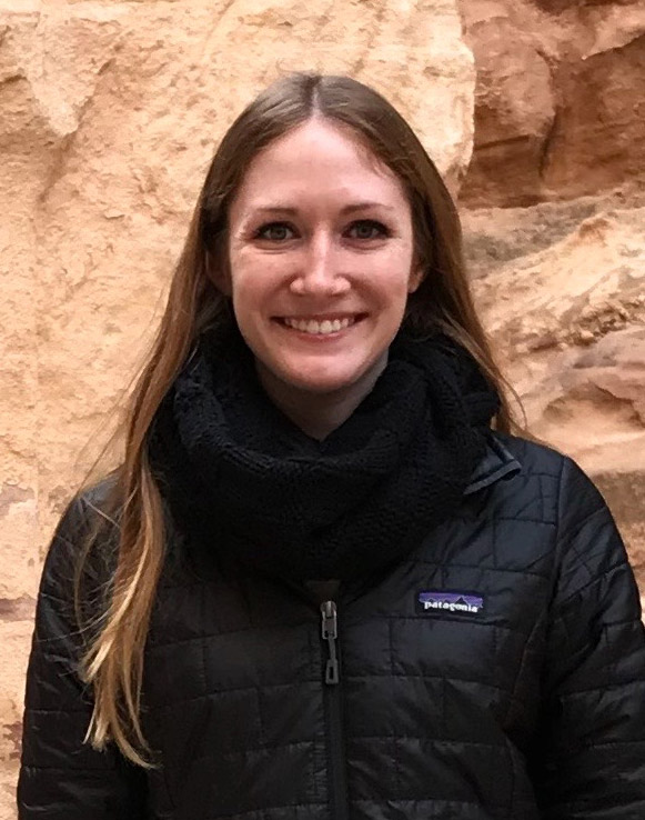 A woman smiling in a black jacket with a scarf, standing outdoors.