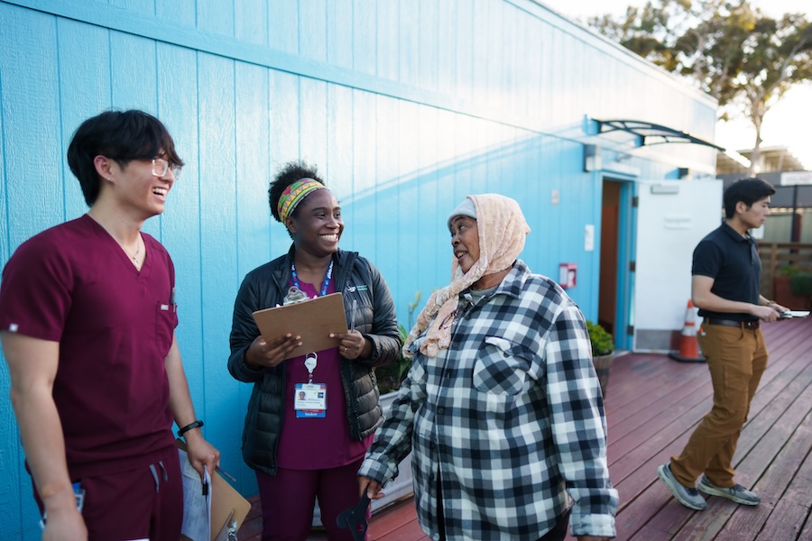 Two healthcare professionals smiling at a woman in a plaid shirt outdoors.
