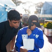 Two men outdoors looking at a white sheet of paper, one man holding an eye patch paddle up to his eye.